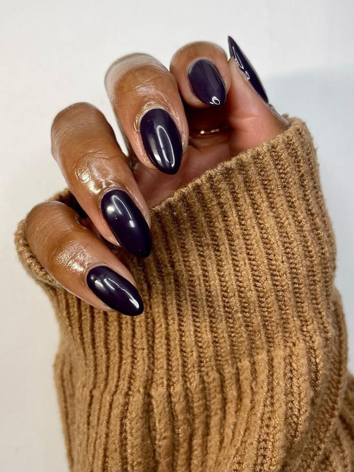 Deep plum almond nails on dark skin, hand in a camel knit sweater, glossy dark purple gel