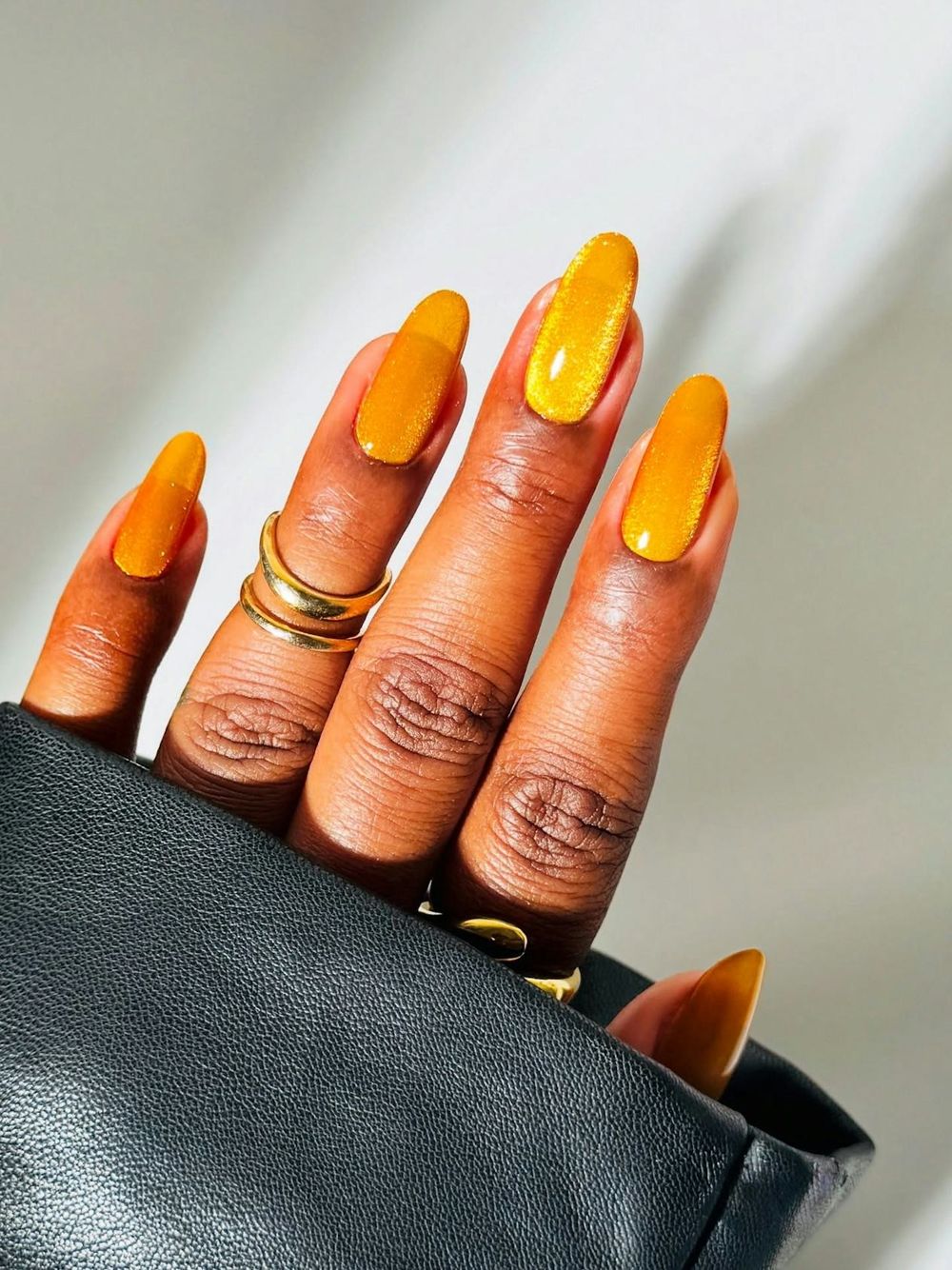 Mustard yellow glossy almond nails on dark skin worn with gold rings and a black leather sleeve