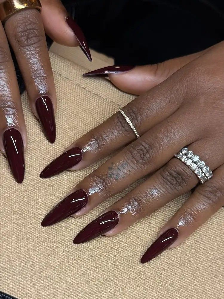 Oxblood stiletto nails on dark skin worn with diamond rings, deep glossy red brown gel