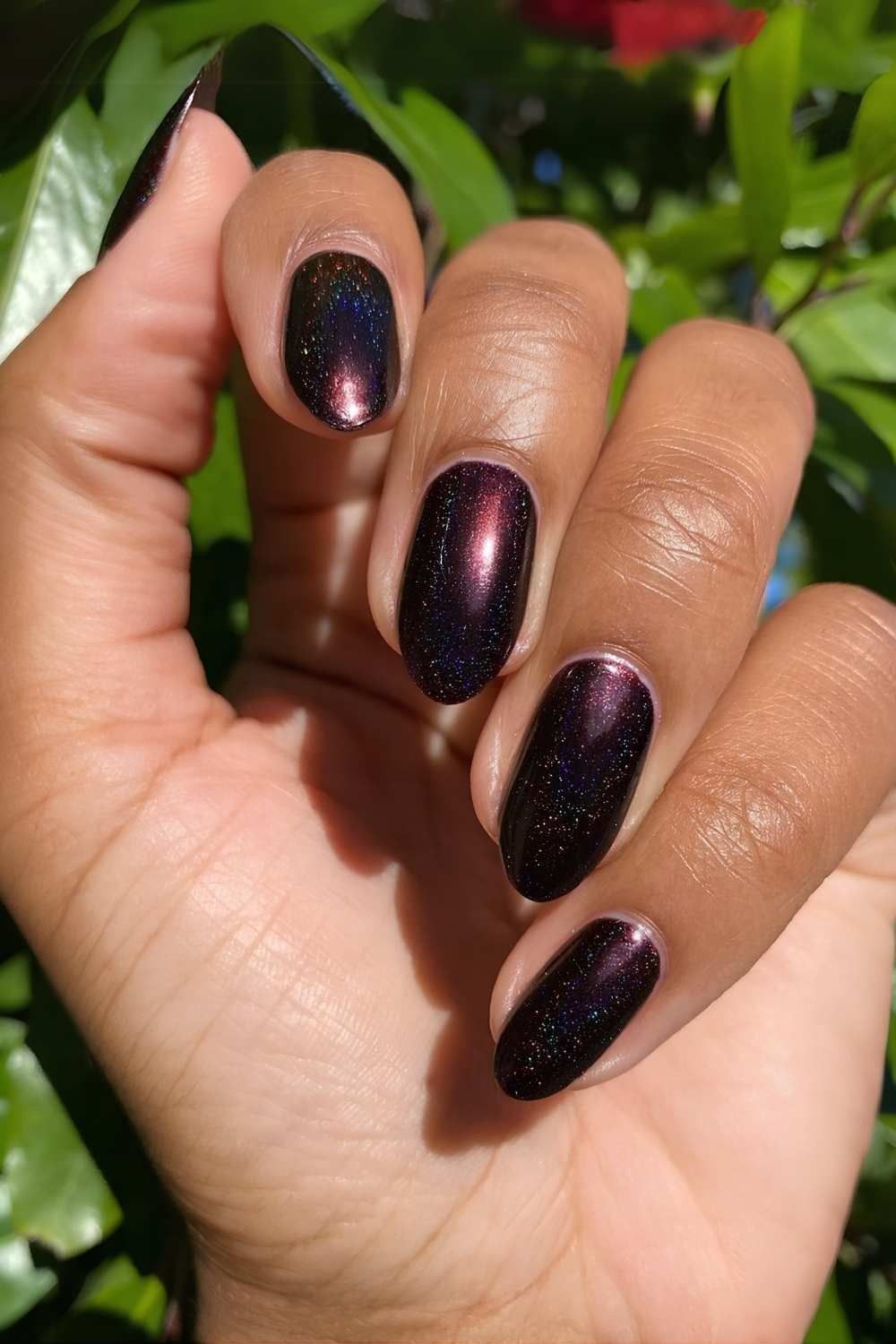Plum galaxy cat eye nails on brown skin photographed outdoors with green leaves, purple shimmer gel