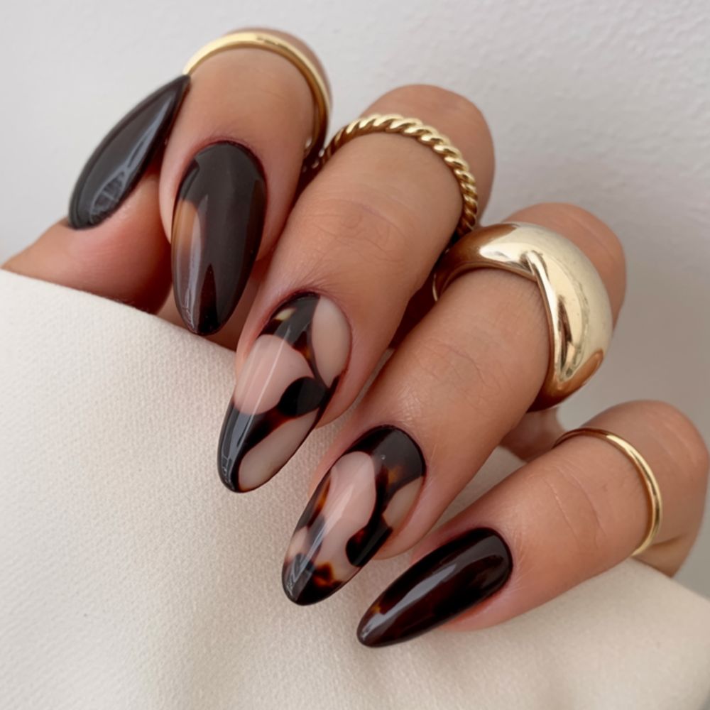 Black and tortoiseshell swirl almond nails, abstract dark swirls over a nude base with chunky gold rings