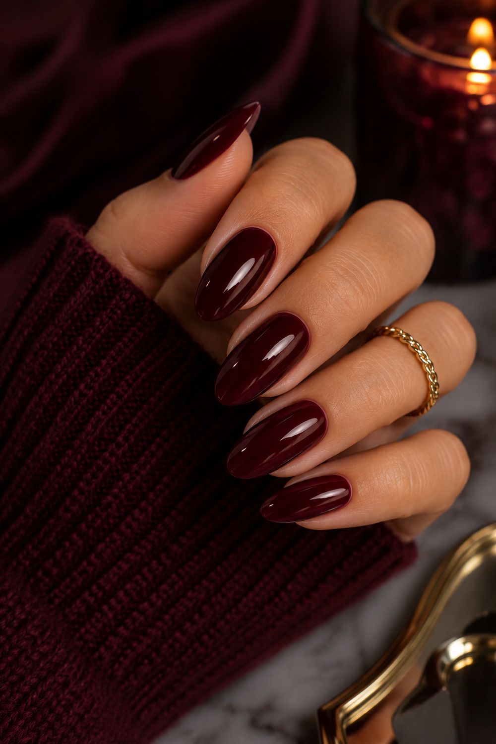 Cozy burgundy nails on a hand in a burgundy knit sweater sleeve, candlelight and fall leaves in the background