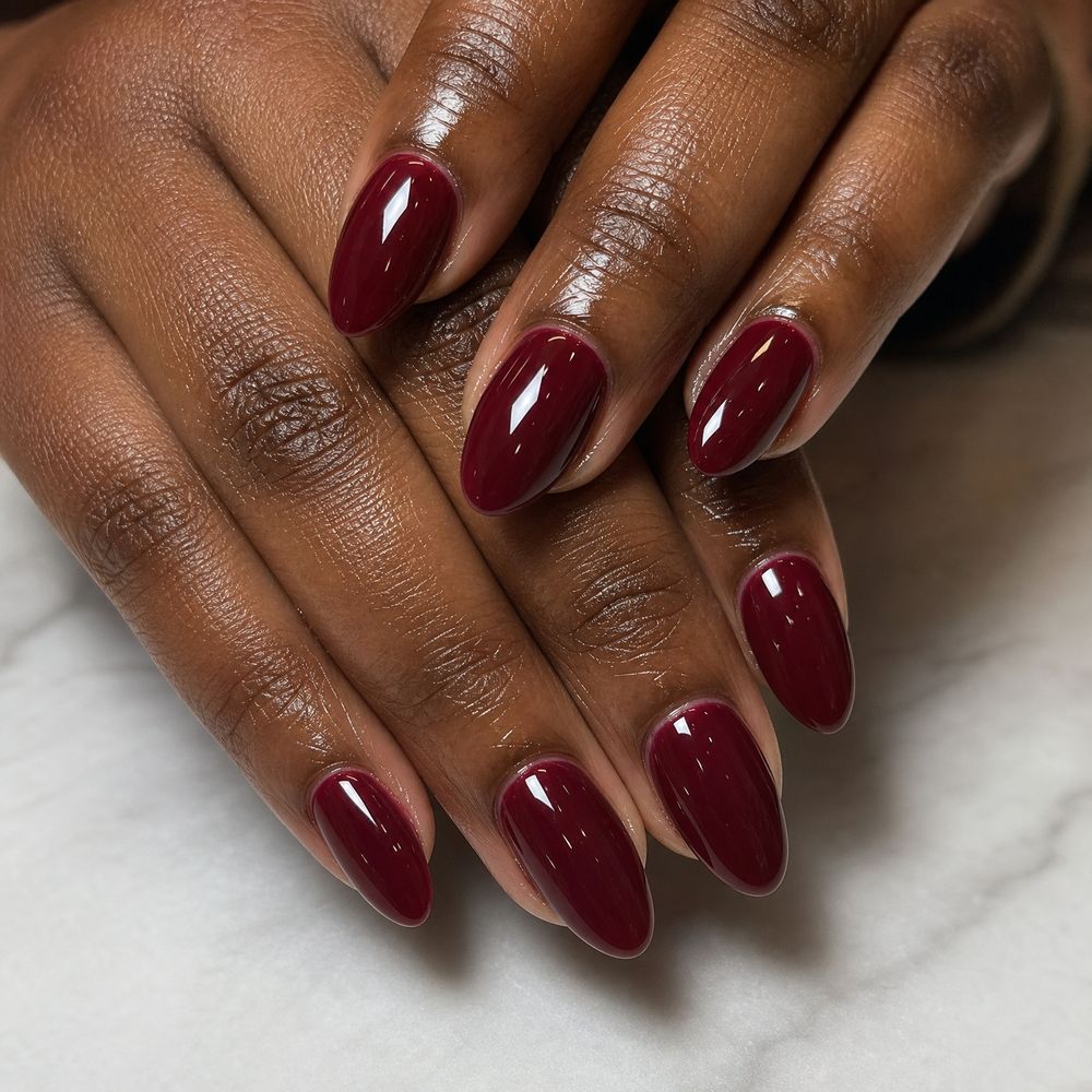 Deep wine almond nails on dark skin, hands crossed on a marble surface, glossy burgundy gel